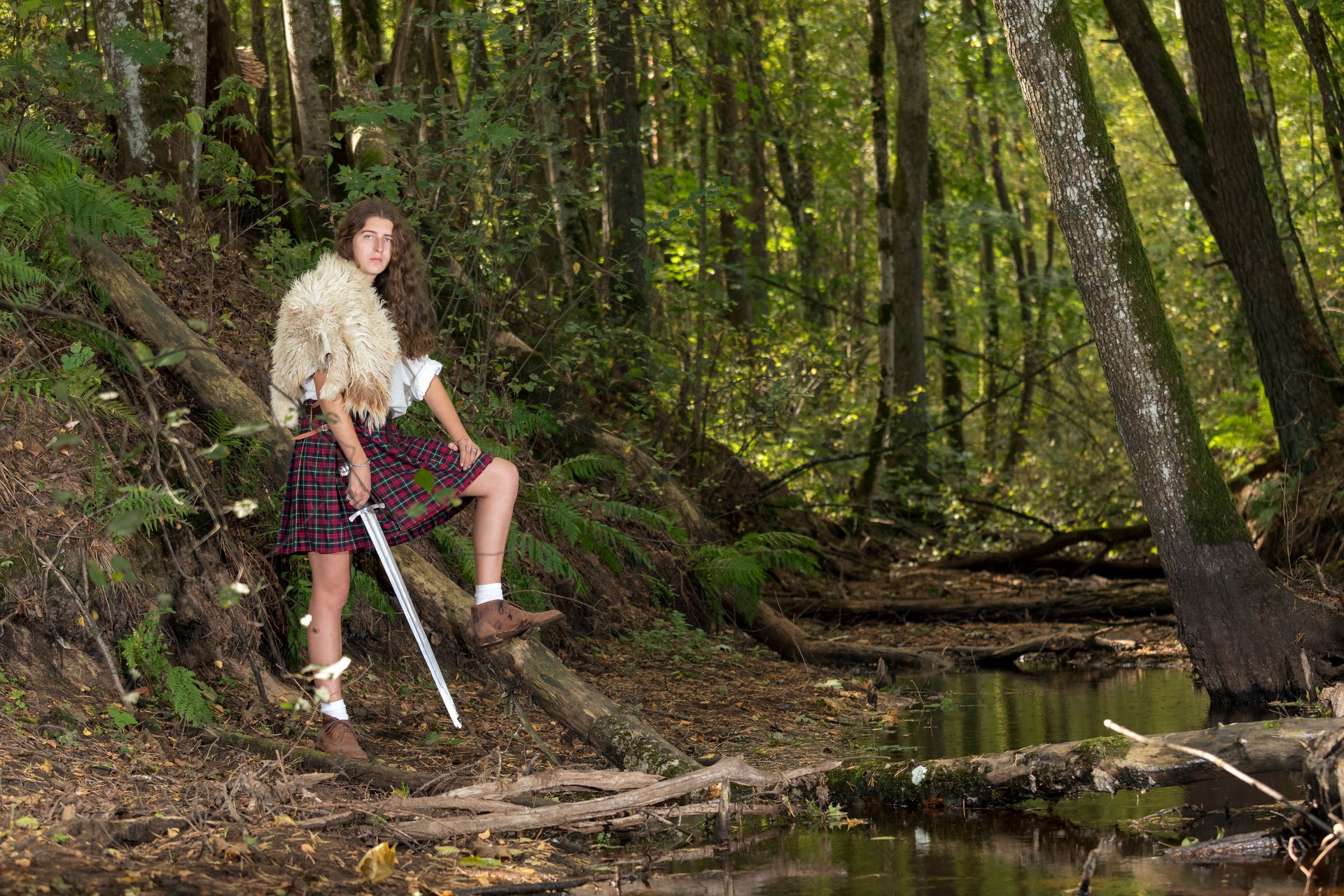 Girl in a Scottish kilt and animal skin on his shoulder posing in forest holding a Viking sword. Eyes facing camera